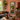 A tall, ornate rectangular mirror with a carved frame hangs on a dusty pink wall in a richly decorated living room. The room features a teal sofa with colorful patterned pillows, a rustic wooden coffee table with a vase of pink flowers, and a carved wooden cabinet with a potted plant.