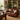 A brown leather loveseat sofa in a styled study or library setting, featuring a wooden desk, brass lamp, bookshelves, and a striped throw pillow.