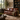 A brown leather loveseat sofa in a styled living room setting, featuring a green throw blanket draped over one side, set against a background with a bookshelf, window, and rug.