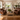 A bright, traditional living room featuring two brown leather club chairs flanking a rustic wooden coffee table on a jute rug, with a cream sofa and large windows in the background.