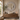 A large round decorative wall mirror with an ornate carved white frame hangs above a rustic wooden floating vanity in a warm beige bathroom. The vanity holds a stone vessel sink, brass faucet hardware, a vase with greenery, soap dispensers, and a lit candle, with a beige towel hanging on the right.