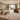 A luxurious, modern bedroom interior featuring a large bed with neutral bedding, a rustic wooden bench, a brown leather armchair with a white throw, and a massive cream shag rug. The room has sage green walls, recessed ceiling lights, and large glass doors opening to a balcony overlooking the ocean.