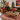 A styled living room interior featuring a carved wooden coffee table in the foreground, a green velvet sofa with patterned cushions, a brown leather armchair, a large patterned area rug, and colorful wall art.